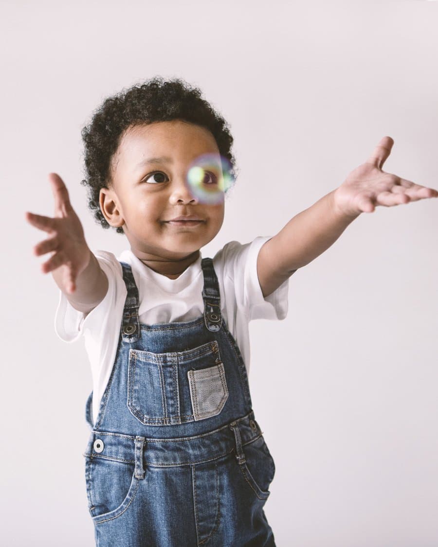 A smiling toddler in denim overalls reaching out for a floating soap bubble